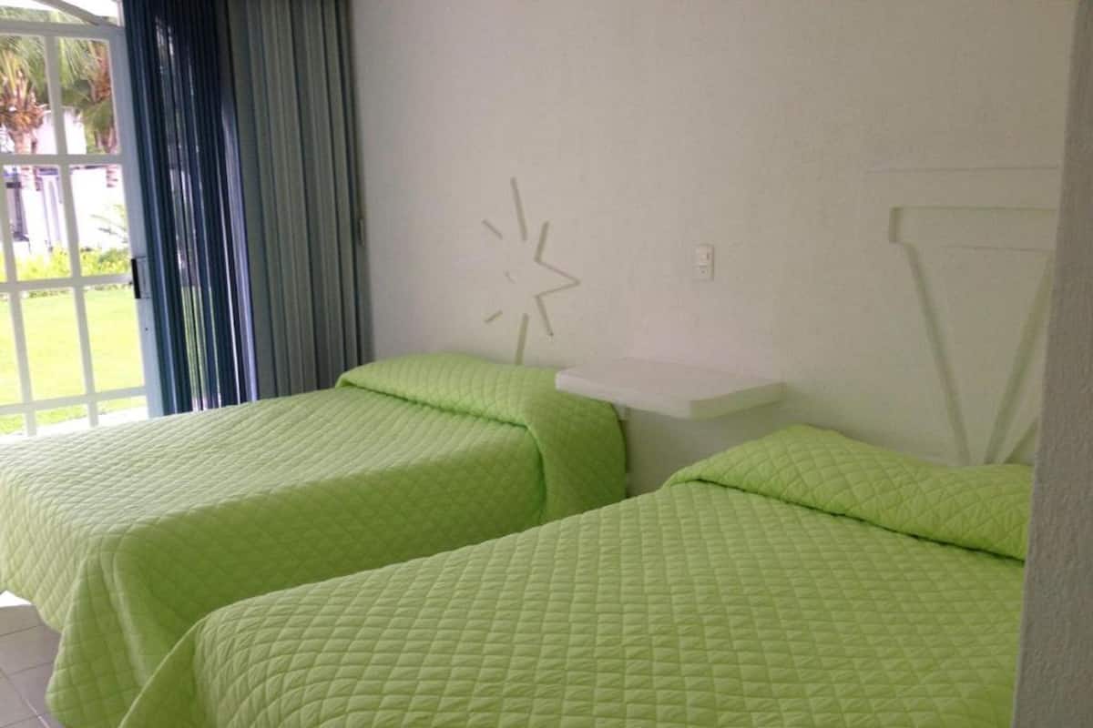 Una habitación con dos camas, cada una con una colcha verde acolchada, una mesita de noche blanca y un reloj de pared.