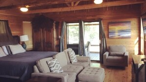 Interior de una cabaña de madera con cama, sofá, sillón, ventanas amplias y un cuadro en la pared.