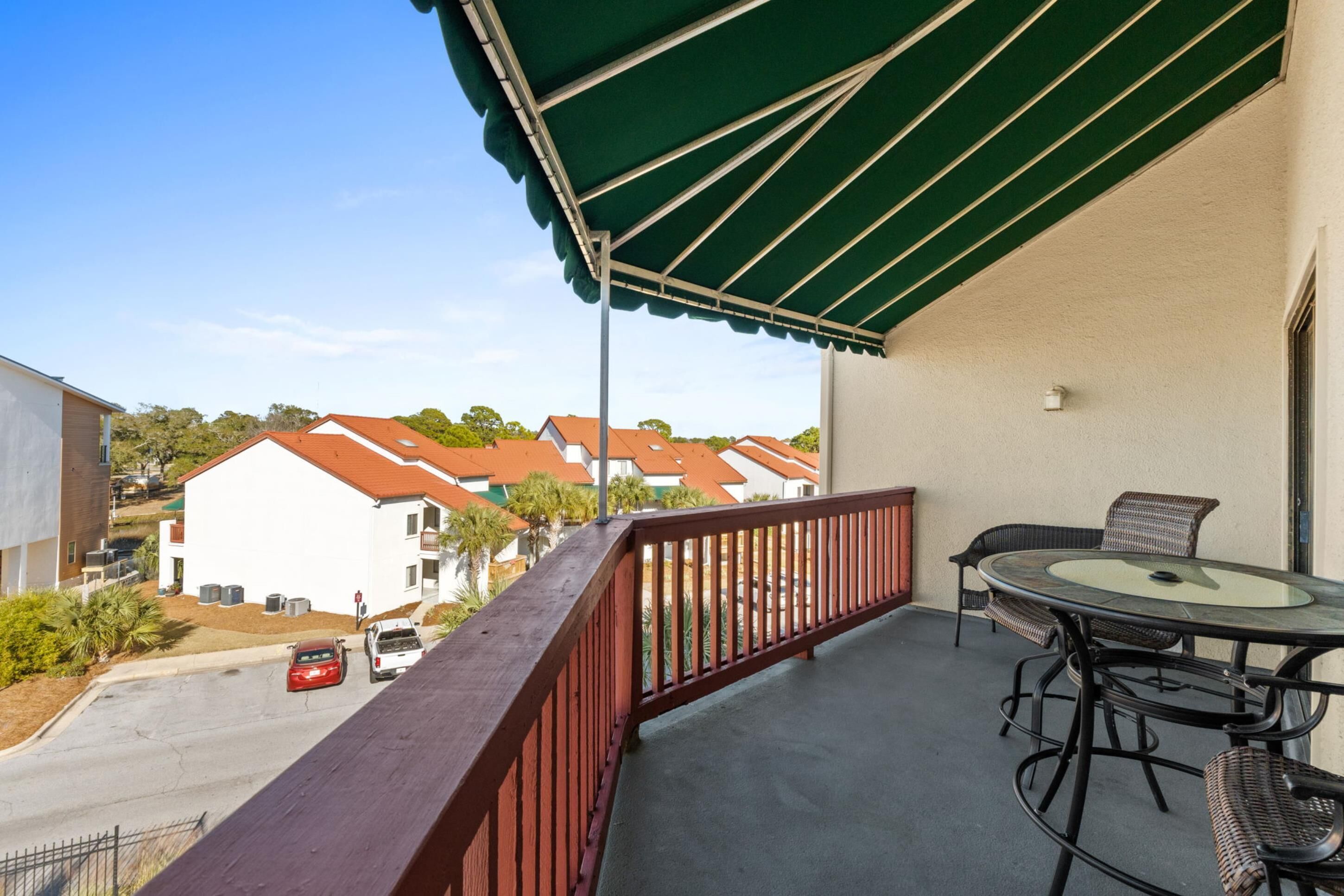 A balcony with a green awning, a round glass table, wicker chairs, and a view of houses with red roofs.