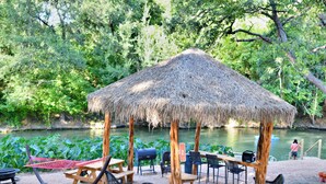 Un pabellón de techo de paja con asientos al aire libre y una parrilla, rodeado de vegetación exuberante y una masa de agua.