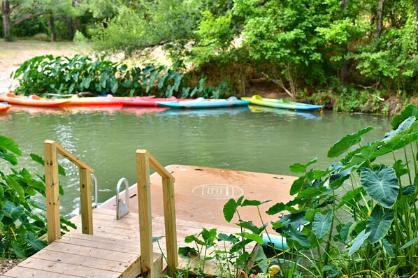 A wooden dock leading to a calm river with colorful kayaks lined up on the shore.