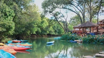 A calm river with colorful boats docked on the left, surrounded by lush greenery and a thatched-roof structure on the right.