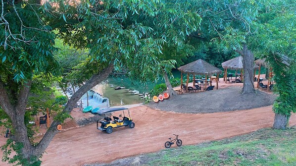 A rustic outdoor setting with a dirt path, a small waterfall, and a thatched-roof gazebo.