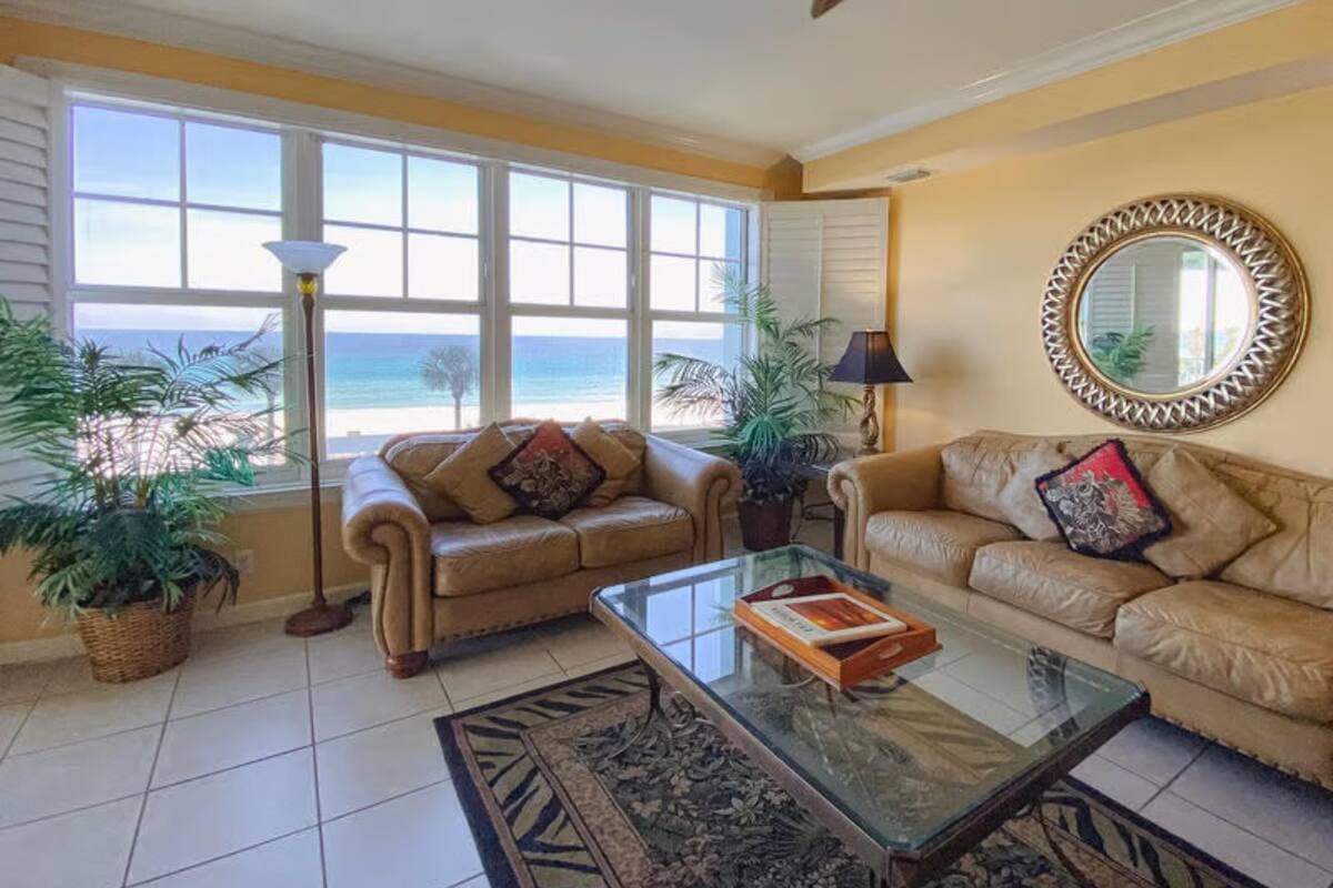 A living room with two sofas facing a large window overlooking the beach. There is a glass coffee table in the center.