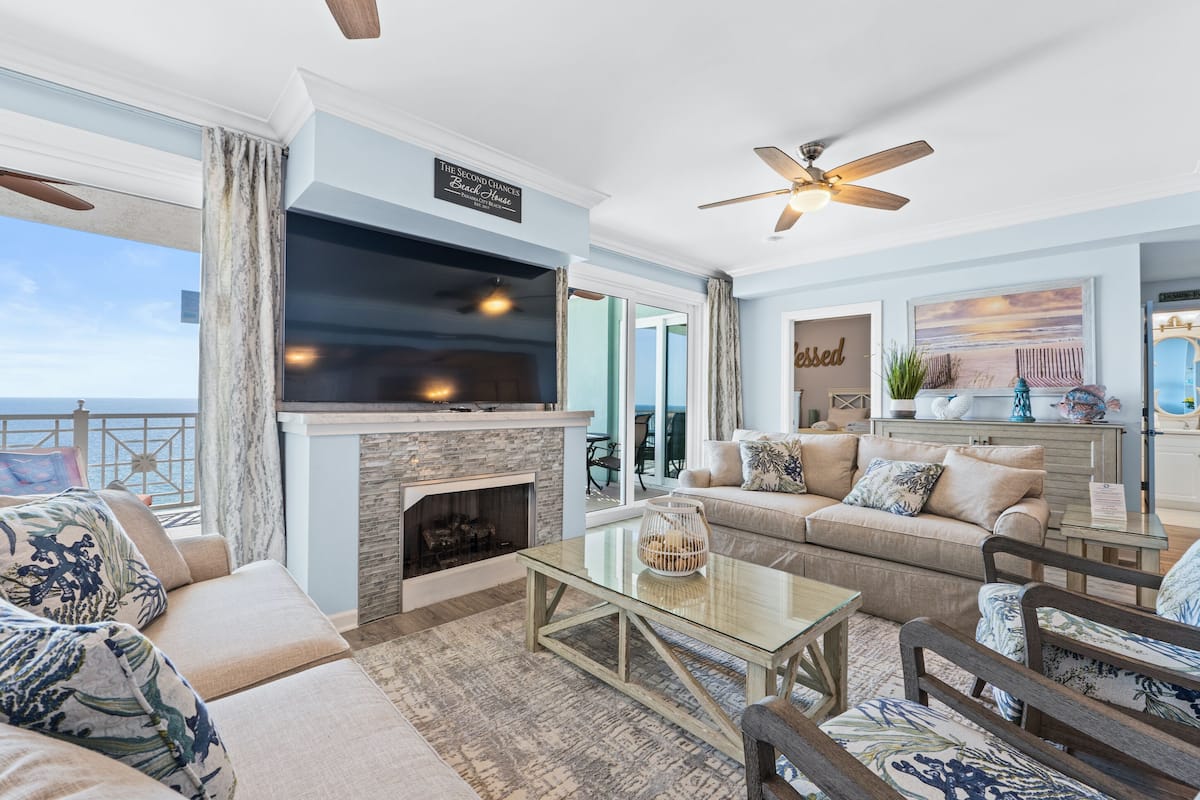 A spacious living room with a fireplace, comfortable seating, a large TV, and a balcony with a view of the sea.