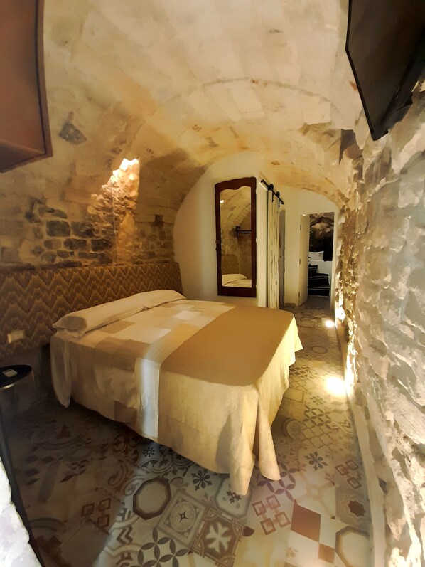 Ein Schlafzimmer mit Steingemäuer, einem Bett mit gemustertem Überwurf und einem Spiegel an der Wand.