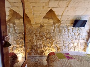 Ein Zimmer mit Steinwänden, einem Bett, einem an der Wand montierten Fernseher und einer Holztür.