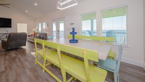 A bright dining area with a wooden table and benches, a television, and a view of the outdoors through large windows.