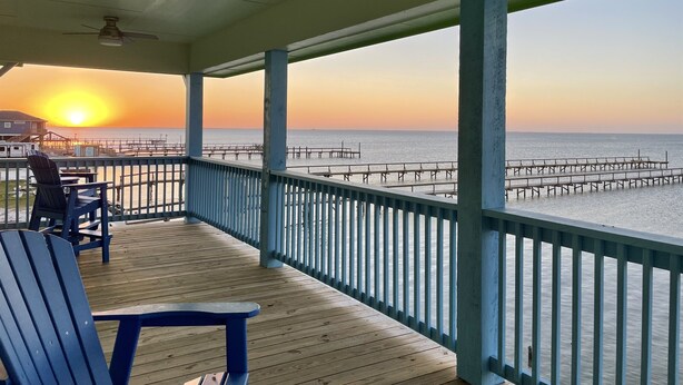 A wooden deck with blue chairs and a table, overlooking a pier extending into the water during sunset.