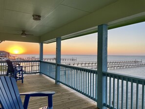 Een houten terras met blauwe stoelen en een tafel, met uitzicht op een pier die in het water uitloopt tijdens de zonsondergang.