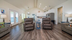 A spacious living room with two brown leather chairs, a coffee table, and a sofa with a colorful pillow. A kitchen is visible in the background with white cabinets and a refrigerator.