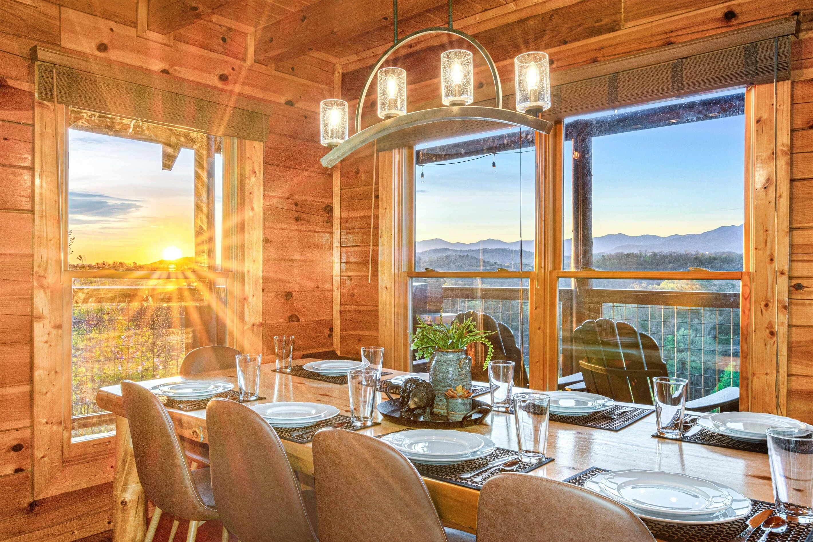 A wooden cabin dining area with a table set for four, large windows, and a view of the mountains.