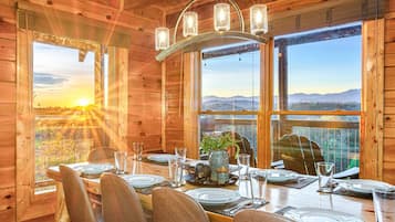 A wooden cabin dining area with a table set for four, large windows, and a view of the mountains.