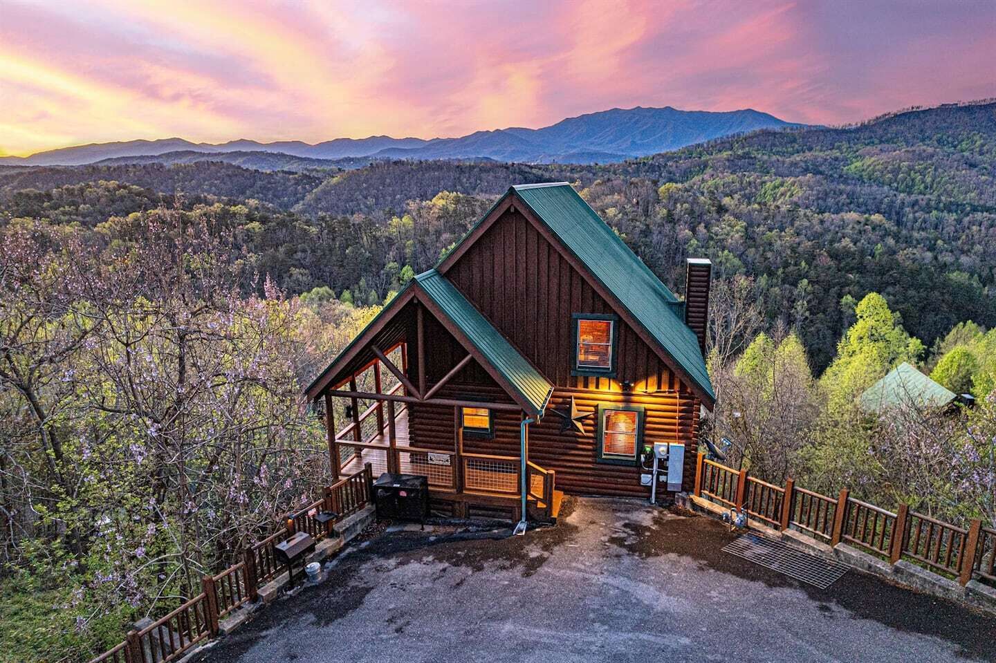A wooden cabin with a green roof, surrounded by a wooden deck and railing, set against a mountainous backdrop.