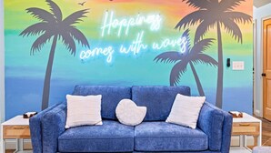 Una habitación con sofá azul, dos almohadas blancas, un letrero de neón que dice 'Hoppings come with waves' y un mural de palmeras y un atardecer.