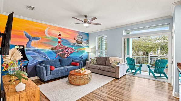 A living room with a mural, two sofas, a coffee table, and a balcony with chairs. - Walk 2 Beach | Newly Redesigned | Kid's Hang Out (Destin)