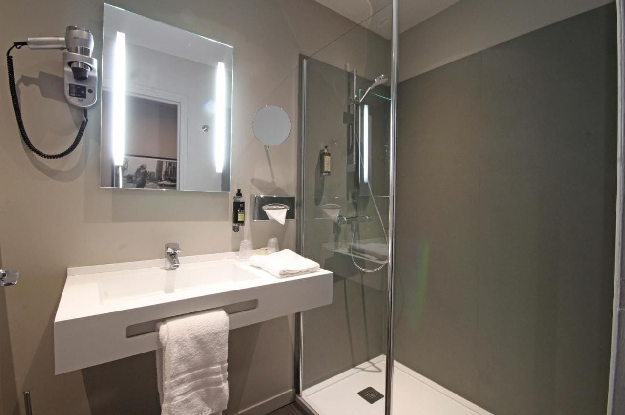 Une salle de bain moderne avec un lavabo blanc, un grand miroir, un sèche-cheveux et une douche à parois de verre.