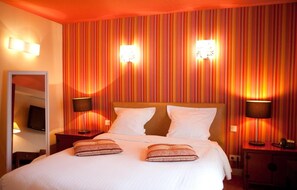 Une chambre d’hôtel avec un mur à rayures, un lit avec du linge de maison blanc, deux lampes de chevet et un miroir sur le mur de gauche.