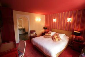 Habitación de hotel con una cama grande, dos lámparas de noche, una silla, una mesita y una puerta que da a un baño.