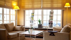 Una sala de estar moderna con un sofá, sillones, una mesa de centro y una planta en maceta. Hay ventanas amplias con persianas y vistas al exterior.