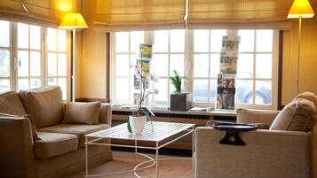Una sala de estar moderna con un sofá, sillones, una mesa de centro y una planta en maceta. Hay ventanas amplias con persianas y vistas al exterior.