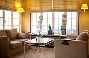 Un salon moderne avec un canapé, des fauteuils, une table basse et une plante en pot. De grandes fenêtres, équipées de stores, offrent une vue sur l’extérieur.
