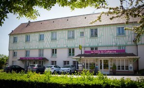 Un bâtiment à deux étages doté d’une façade traditionnelle à colombages, avec une enseigne indiquant « La Table de Catherine » au rez-de-chaussée et « Hôtel Le Faison Doré » à l’étage. Des voitures sont garées devant.