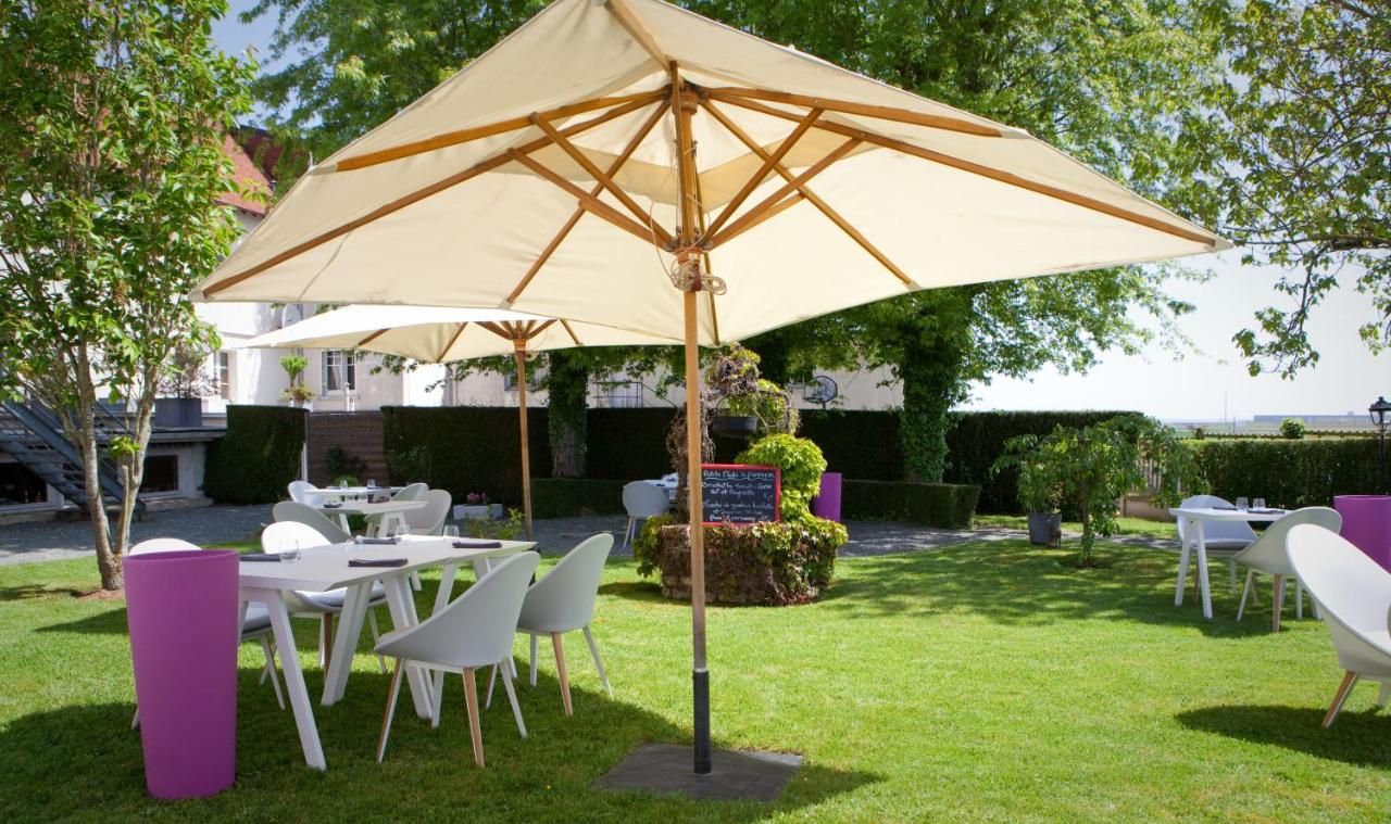 An outdoor seating area with tables and chairs under a large umbrella, surrounded by greenery and a building in the background.