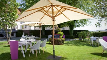 An outdoor seating area with tables and chairs under a large umbrella, surrounded by greenery and a building in the background.
