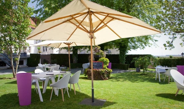 Un espace extérieur aménagé avec des tables et des chaises, protégé par un grand parasol, entouré de verdure et avec un bâtiment en arrière-plan.