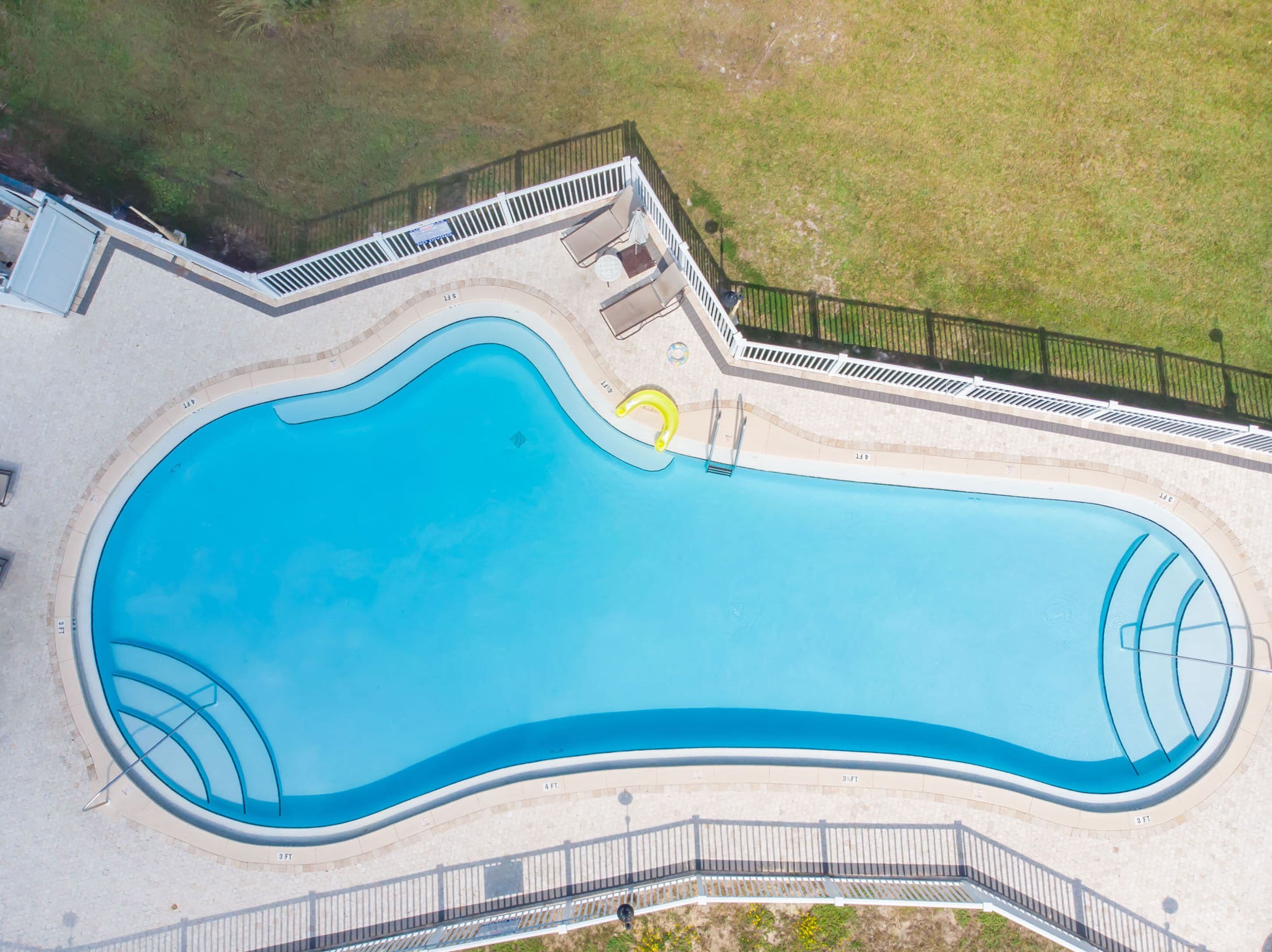 An aerial view of a swimming pool with a curved design, surrounded by a white fence and a small deck area with chairs and a table.