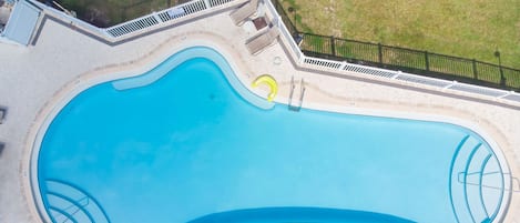 An aerial view of a swimming pool with a curved design, surrounded by a white fence and a small deck area with chairs and a table.