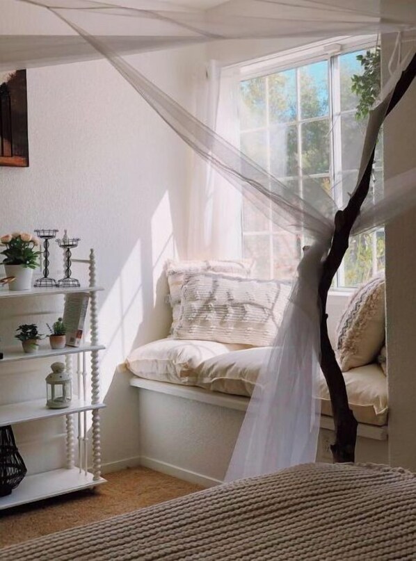 A cozy bedroom with a canopy bed, a white dresser with shelves, and a window with a view of trees.