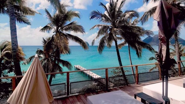 Eine Holzterrasse mit Zelt, Liegestühlen und Blick auf das Meer und Palmen.