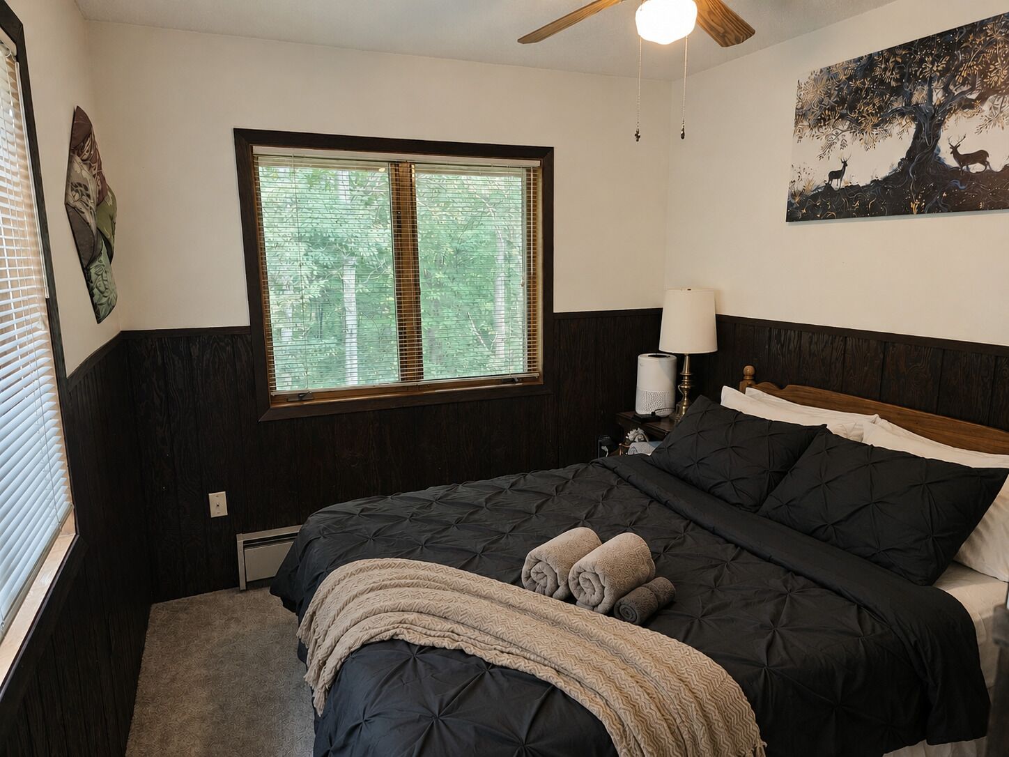 Un dormitorio con una cama grande, una ventana con persianas, un ventilador de techo, una mesita de noche con lámpara y un cuadro colgado en la pared.