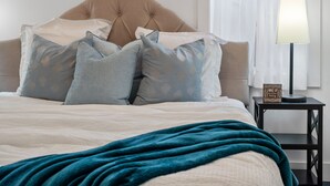 Een keurig opgemaakt bed met een blauwgroene deken, een gebordureerd hoofdbord en meerdere kussens met een bladmotief. Een nachtkastje met een lamp en een bordje met de tekst 'Word Wakker Vandaag' is zichtbaar.