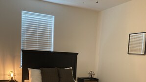 Een slaapkamer met een bed, een plafondventilator, een raam met jaloezieën, een nachtkastje en een vloerkleed.