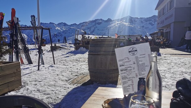 Ein Holztisch mit Speisekarte, einem Glas und einer Flasche, vor einer verschneiten Berglandschaft mit Skiausrüstung.