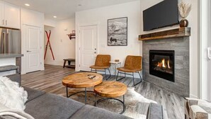 Un salon moderne avec une cheminée, deux tables basses en bois, un canapé gris et un téléviseur à écran plat fixé au-dessus de la cheminée.