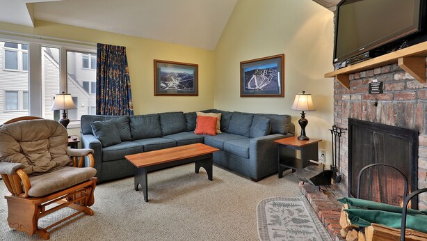 A living room with a sectional sofa, a rocking chair, a fireplace, and framed artwork on the walls. - Beautiful 2BR/2BA Fall Line Condo with Mountain Views on Shuttle Route E2B (Killington)