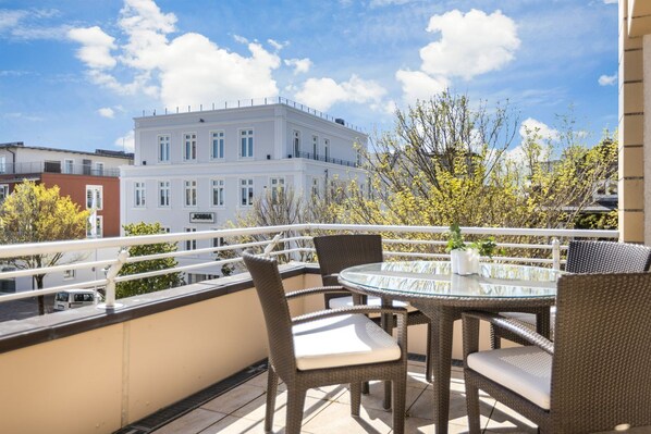 A balcony with wicker chairs and a glass table, overlooking a building with a sign that reads "JONSA".