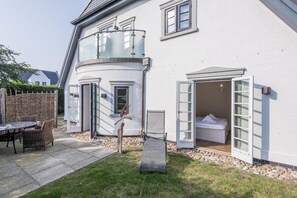 Ein weißes Gebäude mit Balkon, einem Bett mit geöffneten Fensterläden und einem Korbstuhl-Tisch mit Stühlen im Hof.