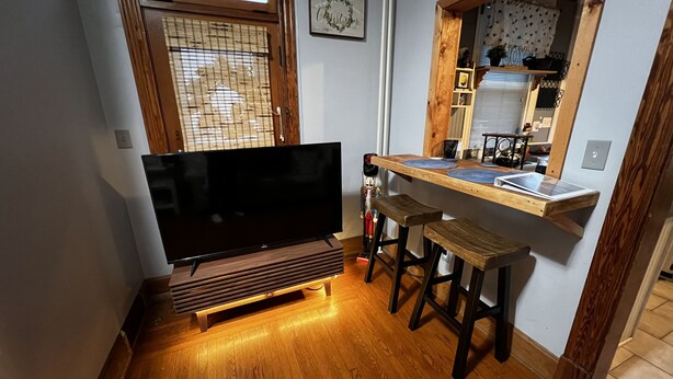 Ein kompakter Wohnbereich mit Flachbildfernseher, Holzbarhocker und einer kleinen Küchenzeile mit Fenster.