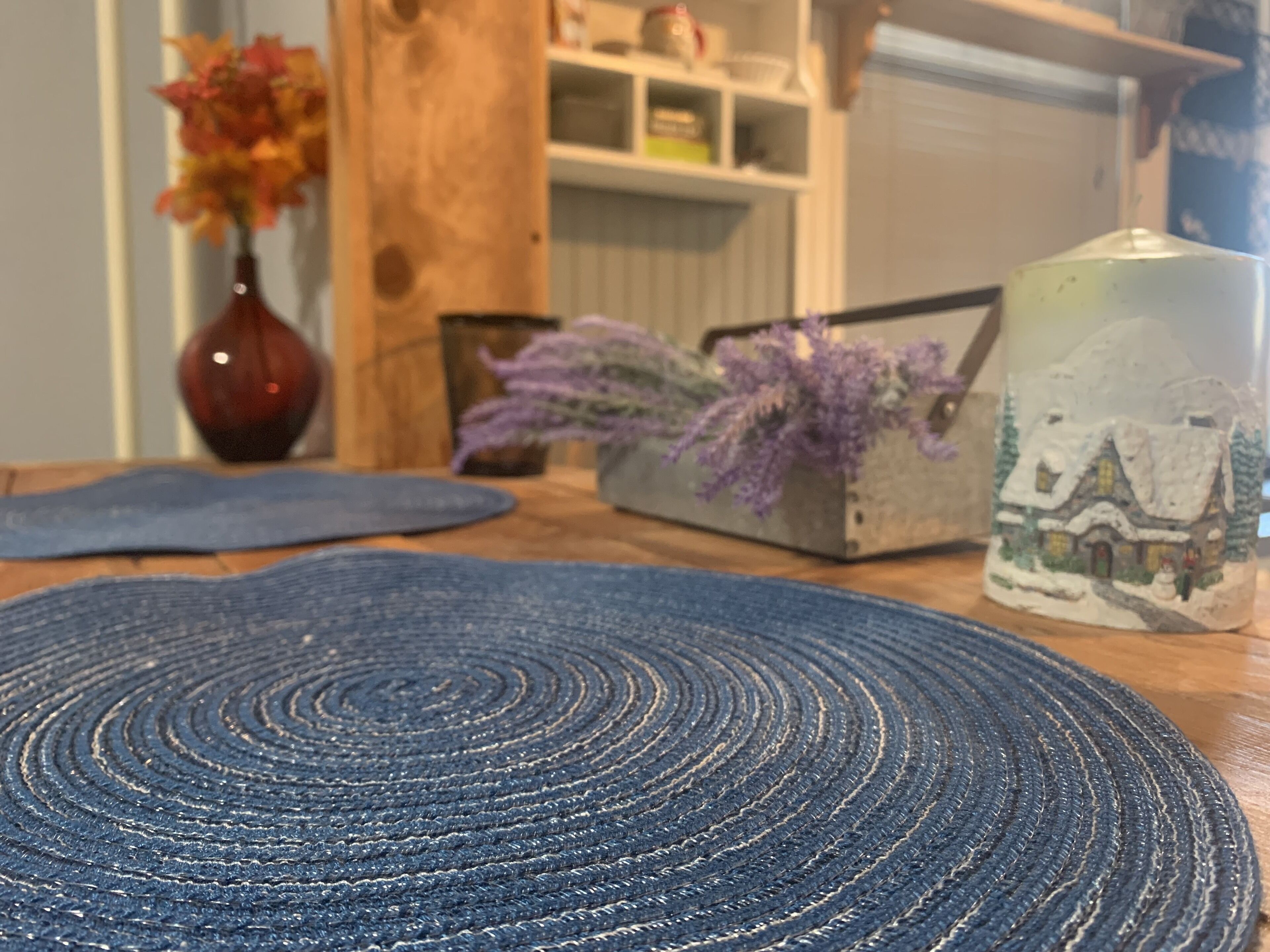 A round blue rug on a wooden table with a vase of flowers and a decorative item in the background.