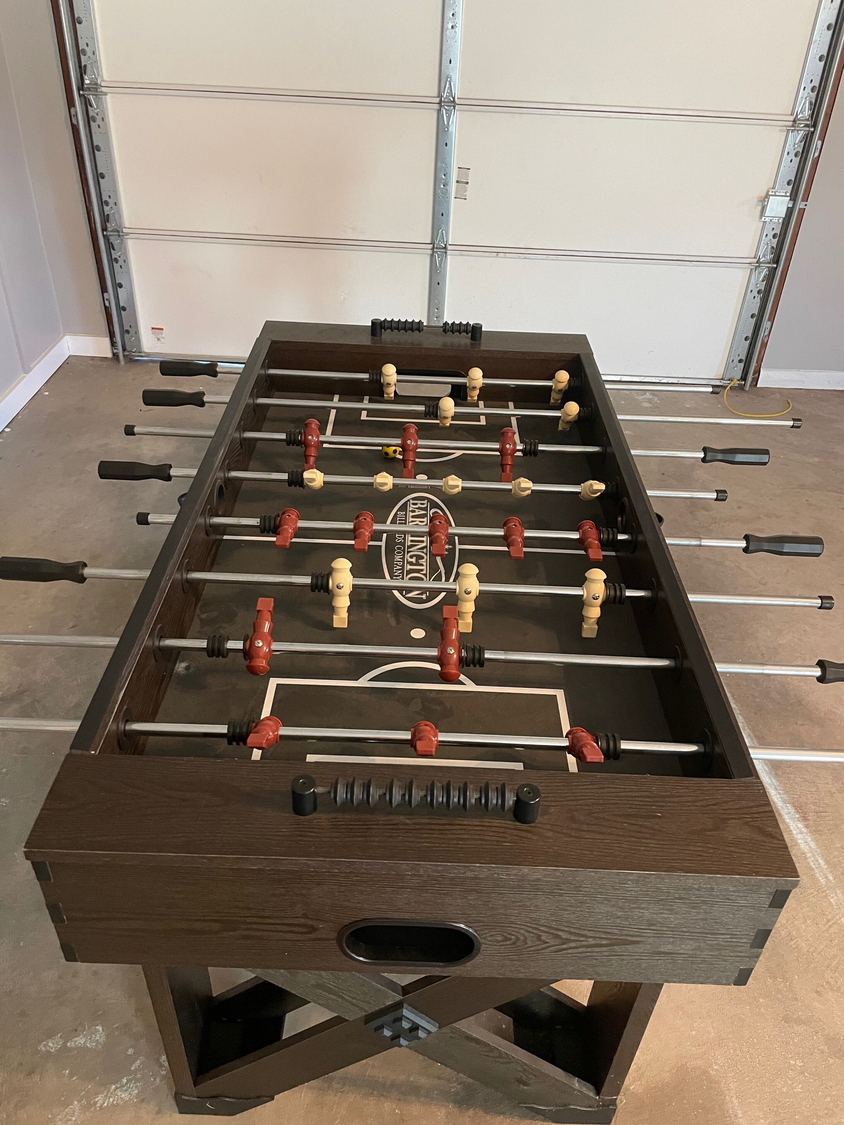 A foosball table with a wooden base and metal rods, set against a garage door.