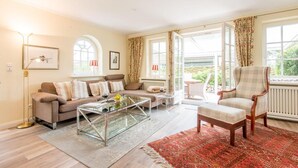 Ein Wohnzimmer mit Sofa, Sessel, Couchtisch und Blick ins Freie durch große Fenster.