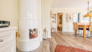 Ein modernes Wohnzimmer mit einem weißen, zylindrischen Gegenstand, möglicherweise einer Heizung, einem Essbereich mit einem Holztisch und Stühlen sowie einem roten Teppich mit Muster.