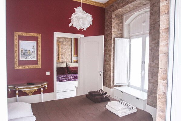A bedroom with a bed, a bedside table, a lamp, a window, and a door leading to another room.