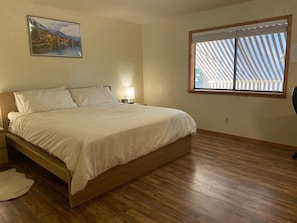 A bedroom with a wooden bed, a nightstand with a lamp, a painting on the wall, and a window with blinds.