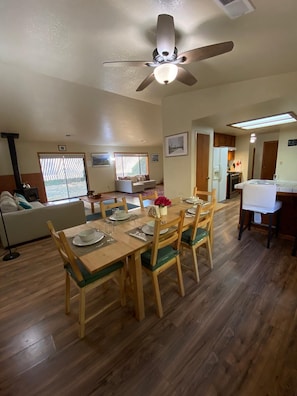 A dining area with a wooden table set for four, a ceiling fan, and a kitchen in the background.